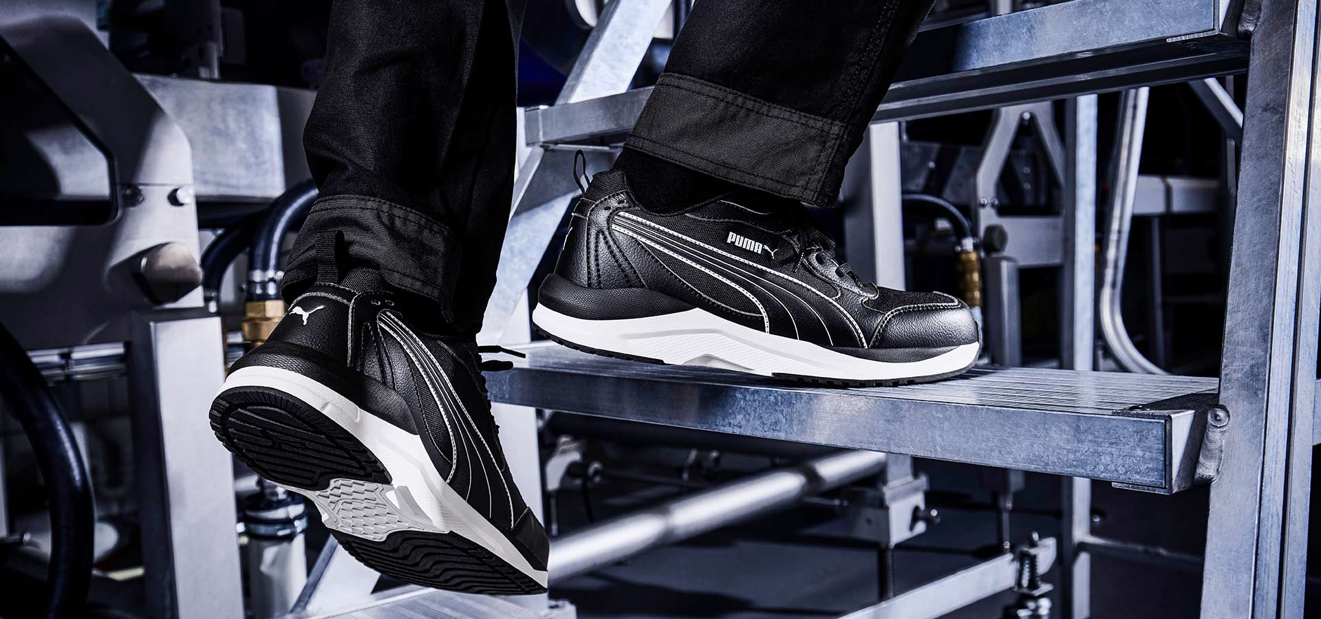 Schwarzer Puma Sicherheitsschuh SHIFT BLK/WHITE LOW S3S FO HRO SR mit weißer Sohle, getragen auf einer Metalltreppe in industrieller Umgebung.