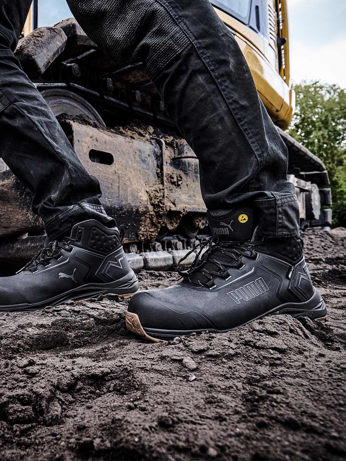 Mobile Ansicht: PUMA SAFETY FUSION CTX MID Sicherheitsschuh S7S ESD in Anthrazit, getragen von einer Person auf schlammigem Untergrund vor einer Baumaschine mit Kettenantrieb im Außenbereich