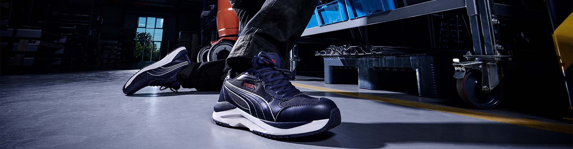 Sicherheitsschuh SHIFT BLUE LOW S1PS FO HRO SR in Dunkelblau mit weißen und roten Akzenten, getragen in einer Werkhalle auf grauem Industrieboden.