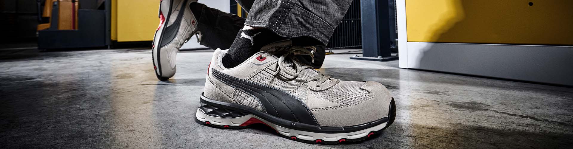 Grauer Sicherheitsschuh VANTI GREY LOW S1PS ESD FO HRO SR von Puma mit schwarzem Streifendetail, getragen auf Werkstattboden vor gelbem Maschinengehäuse.