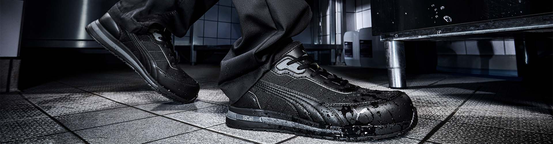 Schwarzer Sicherheitsschuh EPIC BLK LOW S3S im Einsatz auf nassem Fliesenboden, mit wasserabweisendem Obermaterial und rutschfester Sohle.