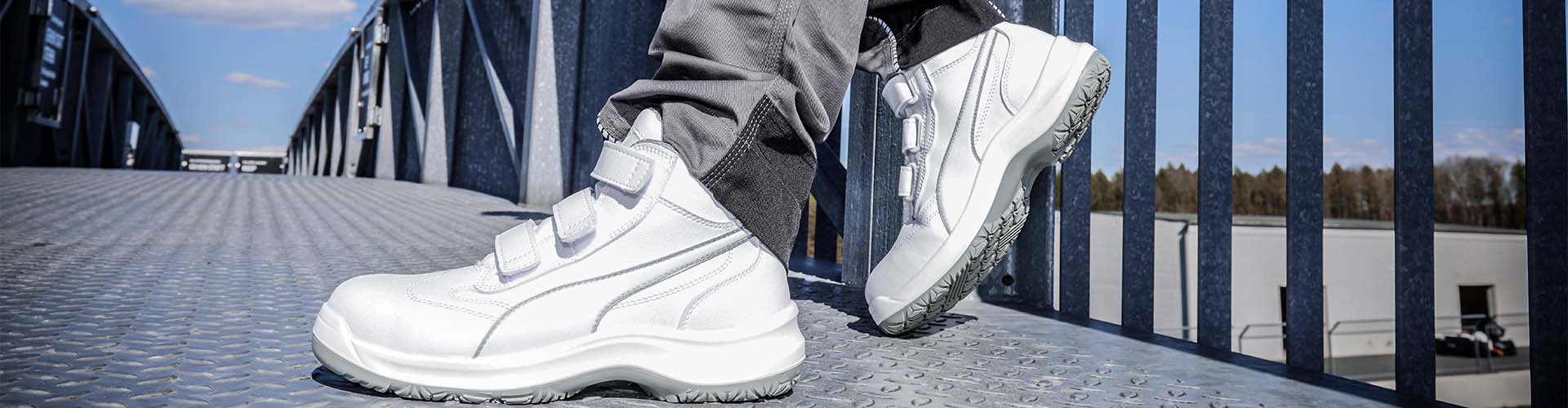 Weißer Sicherheitsschuh ABSOLUTE MID S2 SRC mit Klettverschluss, getragen auf einer Metallplattform im Außenbereich bei Sonnenschein.