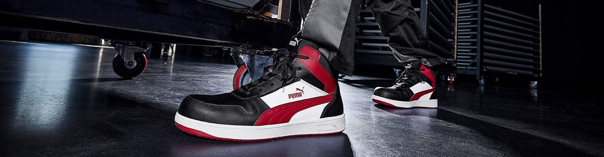 Schwarz-rot-weißer PUMA SAFETY ICONIC LOW Sicherheitsschuh S1PL ESD mit Schnürung und flacher Sohle, am Fuß getragen auf dunklem Boden neben einem rollbaren Metallgestell.