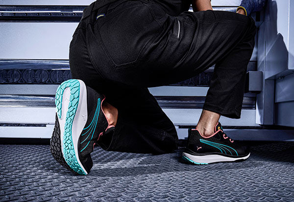 Schwarze PUMA Sicherheitsschuhe mit türkisfarbenen Details und rutschfester Sohle beim Knien auf einem geriffelten Industrieboden.