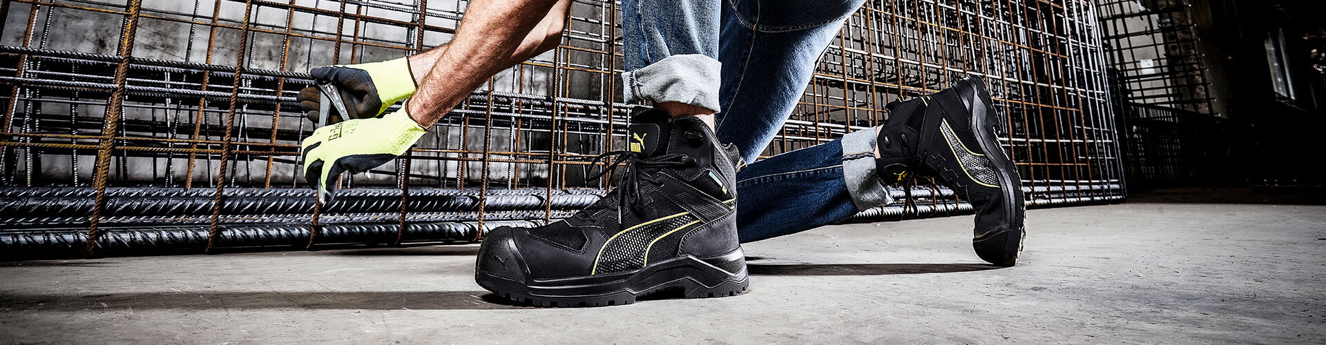 Schwarzer PUMA SAFETY ROCK HD CTX MID Sicherheitsschuh S7S mit gelben Details und profilierter Sohle, am Fuß getragen in kniender Position auf Baustellenboden mit Stahlgittern im Hintergrund.