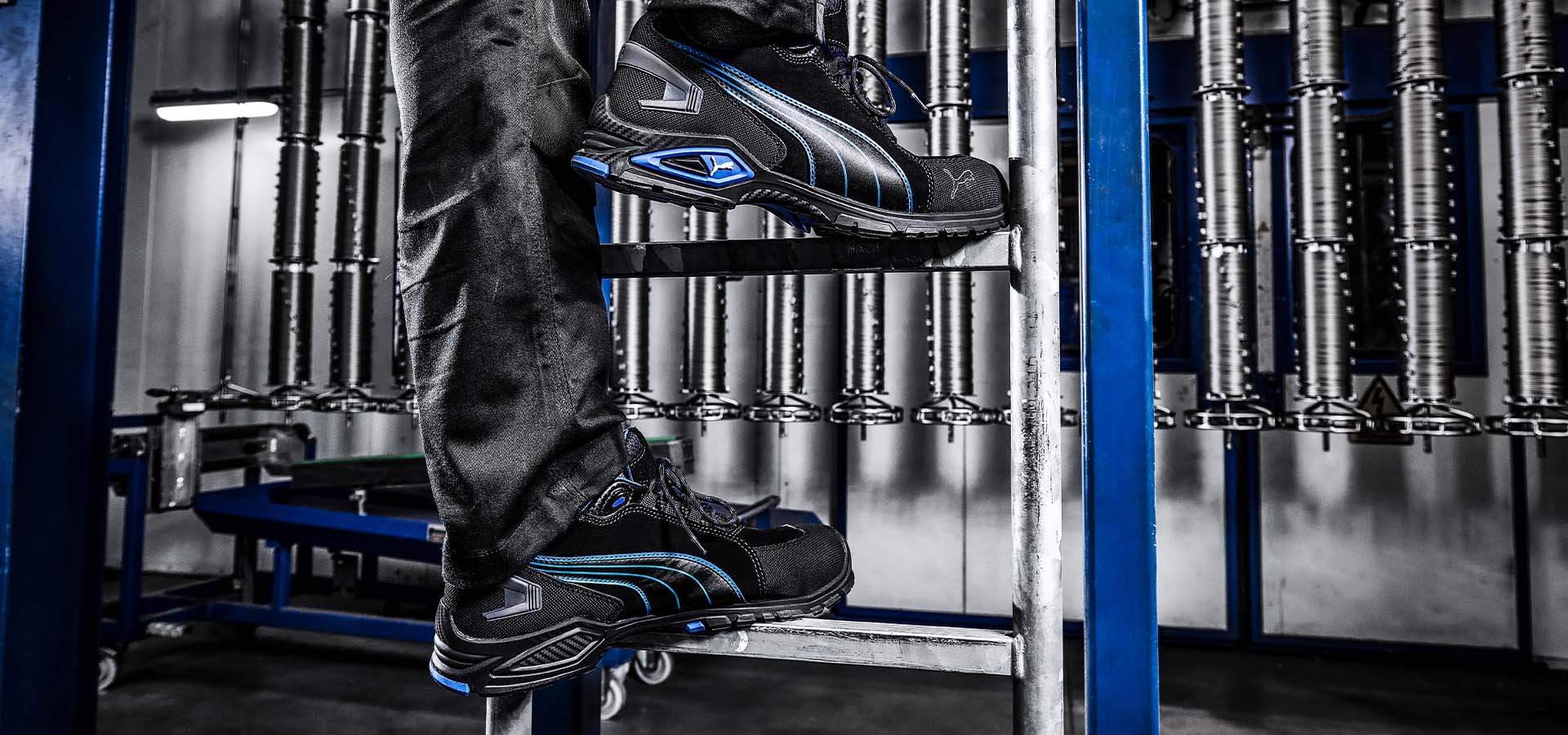 Schwarzer Puma Sicherheitsschuh RIO BLACK MID S3L FO SR mit blauen Details, getragen auf einer Metallleiter in industrieller Umgebung.