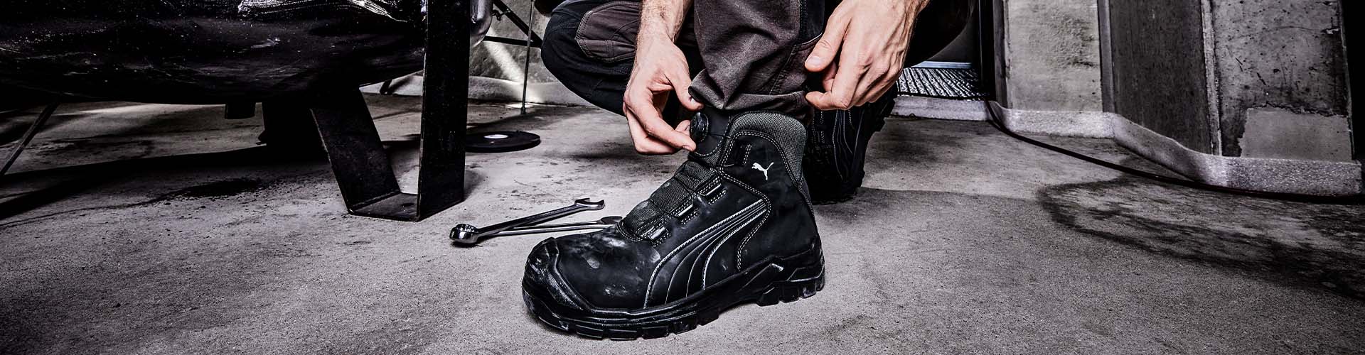 Schwarzer Sicherheitsschuh CASCADES DISC MID S3S FO CI HI HRO SR von Puma, getragen bei der Arbeit auf Betonboden mit Werkzeug im Hintergrund.