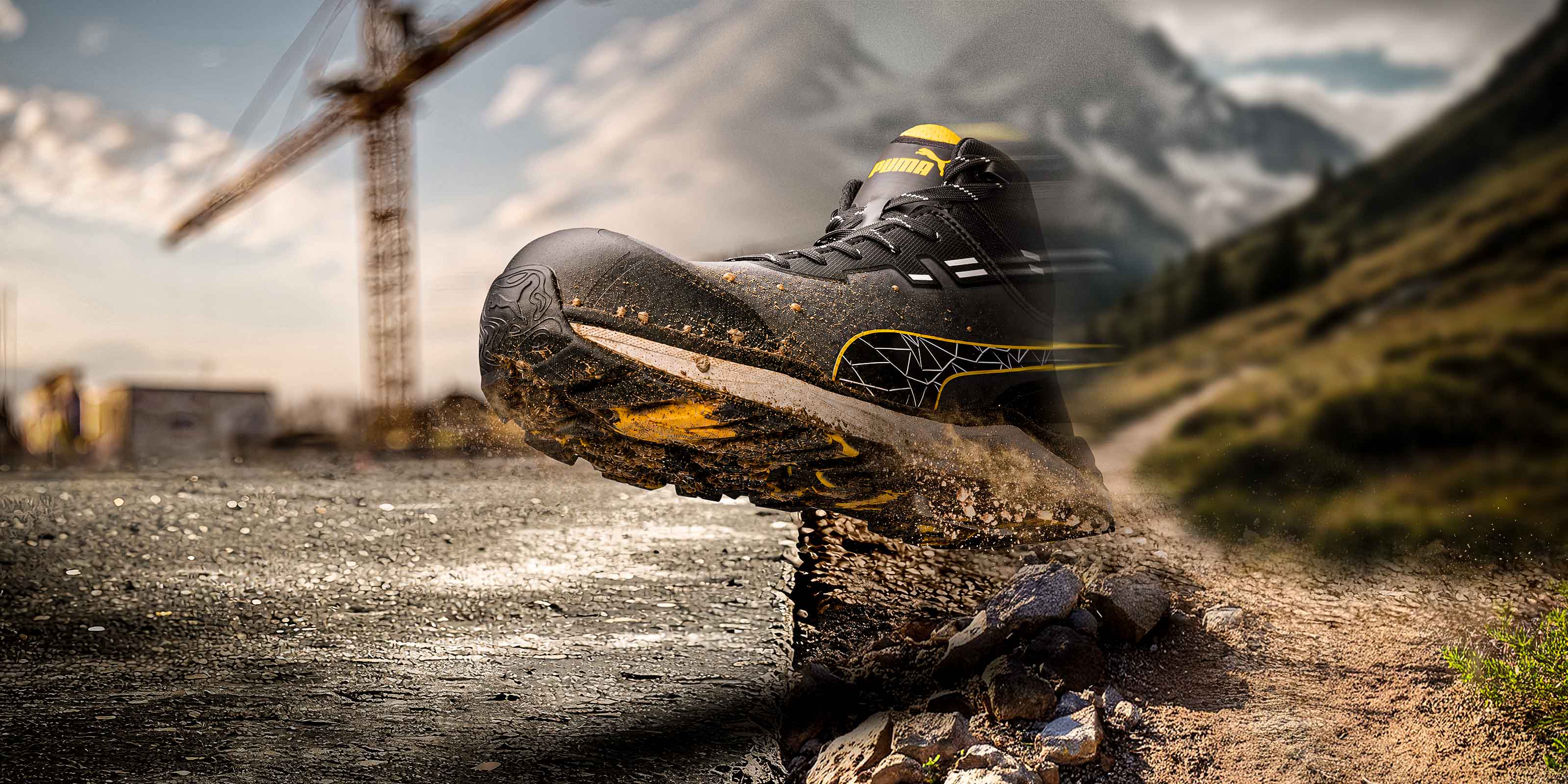PUMA SAFETY XPLORE MID safety shoe in charcoal gray with yellow accents, shown in motion as the wearer steps onto a rocky surface with swirling dirt, against a split-screen backdrop featuring a construction site with a crane and a mountain landscape