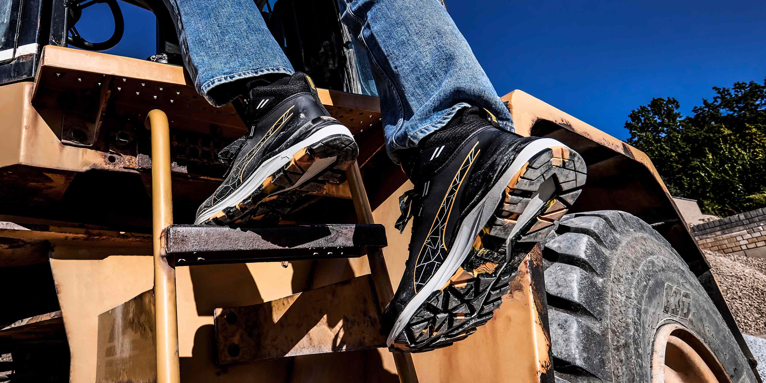 PUMA SAFETY XPLORE MID Sicherheitsschuh S3S ESD in Anthrazit mit gelben Akzenten, getragen von einer Person beim Aufstieg über eine Metallleiter an einer Baumaschine, mit sichtbarer profilierter Laufsohle