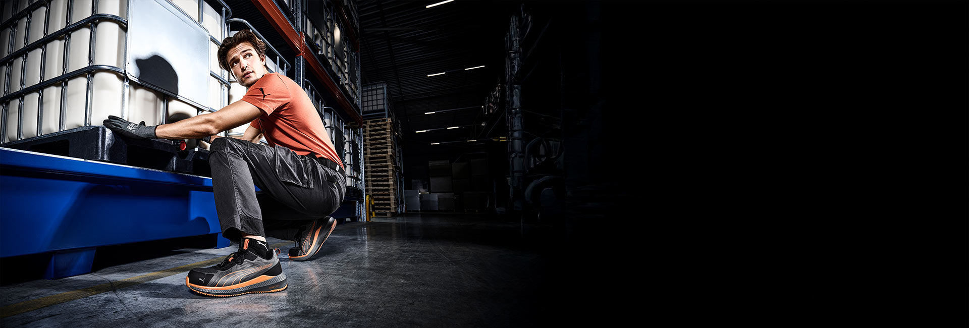 PUMA Sicherheitsschuhe in Schwarz-Orange beim Arbeiten in einer Lagerhalle, getragen von einer knienden Person neben einem großen Industriebehälter.