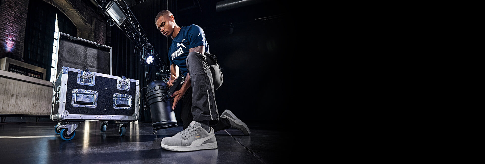 Grauer PUMA SAFETY ICONIC LOW Sicherheitsschuh S1PL ESD mit weißer Sohle und Schnürung, am Fuß getragen von einem Mann beim Arbeiten mit Lichttechnik auf dunklem Boden.