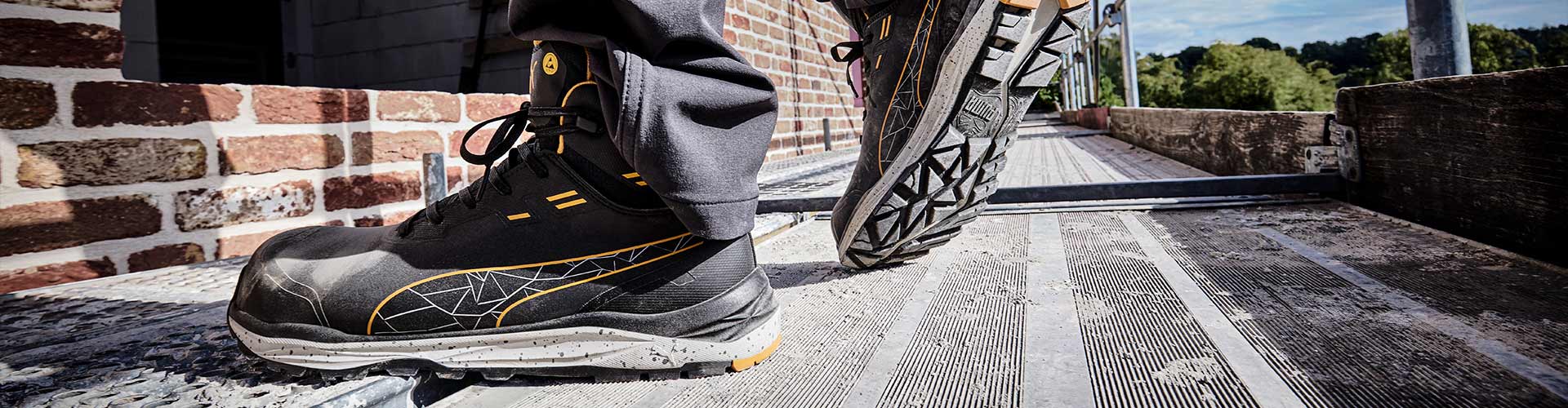 PUMA SAFETY XPLORE MID Sicherheitsschuh S3S ESD in Anthrazit mit gelben Akzenten, getragen von einer Person beim Gehen auf einem Gerüstboden neben einer Ziegelwand im Außenbereich