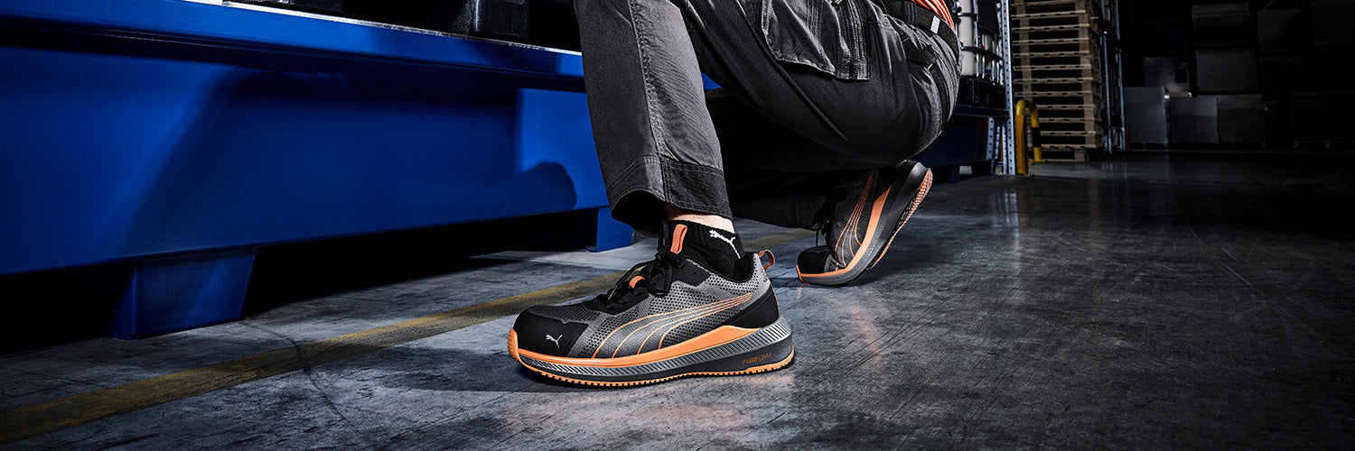 PUMA SAFETY SLIDE LOW Sicherheitsschuh S1PS ESD in Schwarz mit grauen Flächen und auffälligen orangefarbenen Akzenten, leichter sportlicher Sneaker-Stil mit strapazierfähiger Schutzkappe, getragen in industrieller Umgebung.
