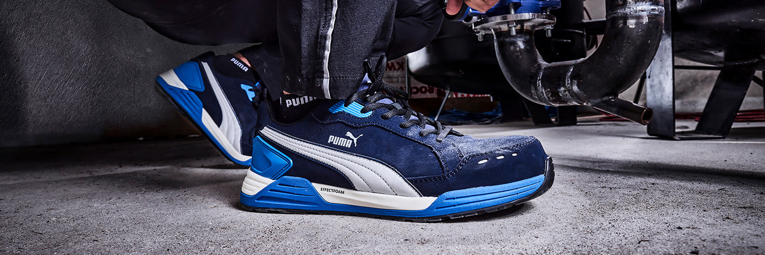 Blau-grauer PUMA SAFETY AIRTWIST LOW Sicherheitsschuh S3 ESD mit weißer Sohle und Schnürung, am Fuß getragen bei Arbeiten auf Betonboden neben Rohrleitungen.