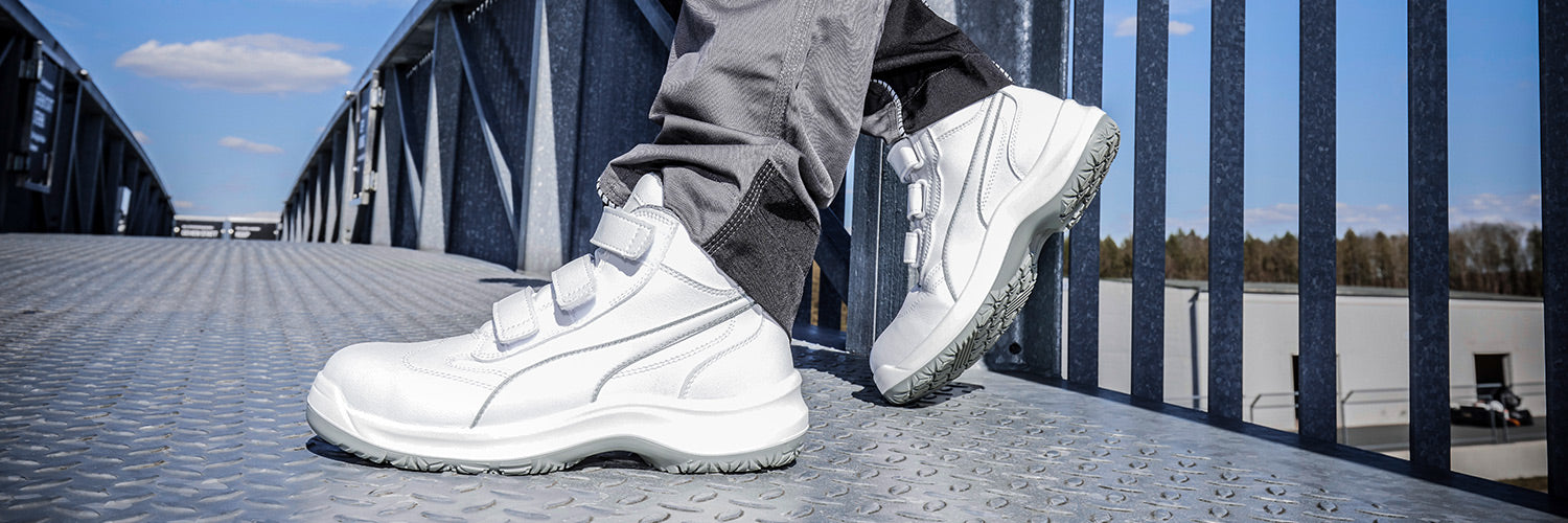 PUMA SAFETY ABSOLUTE MID Sicherheitsschuh S2 in Weiß mit Klettverschluss, getragen von einer Person auf Metallsteg im Freien.