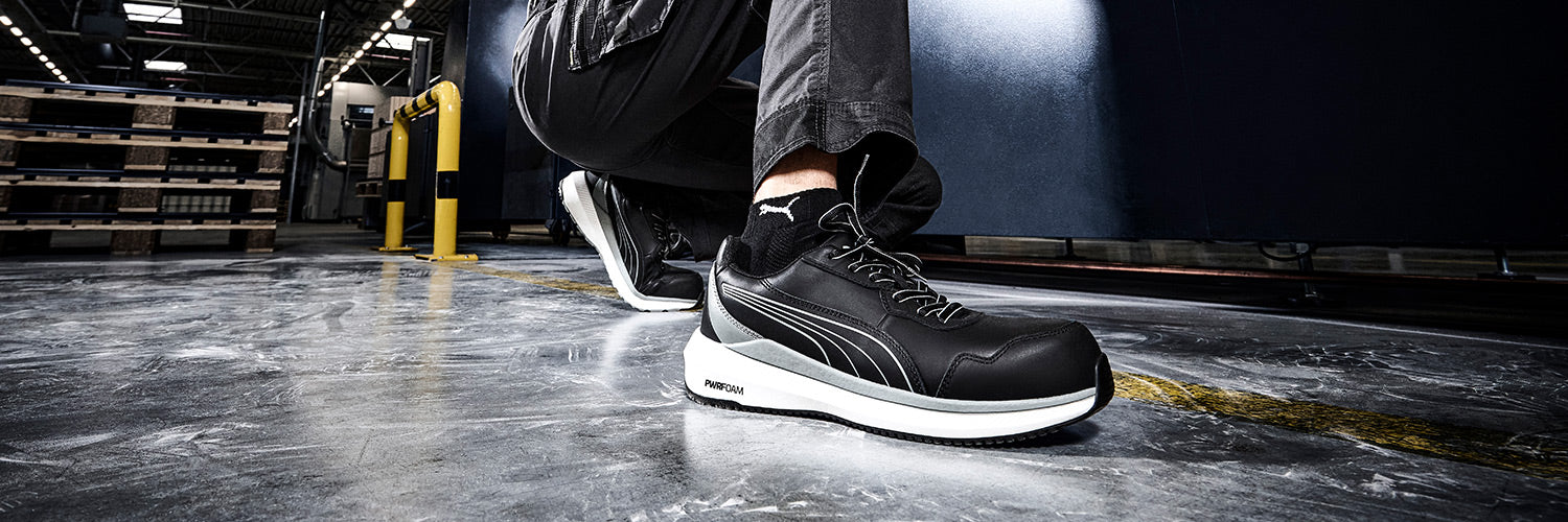 Schwarzer PUMA SAFETY ZOOM LOW Sicherheitsschuh S3S ESD mit weißer Sohle und Schnürung, am Fuß getragen auf glänzendem Industrieboden mit Paletten im Hintergrund.