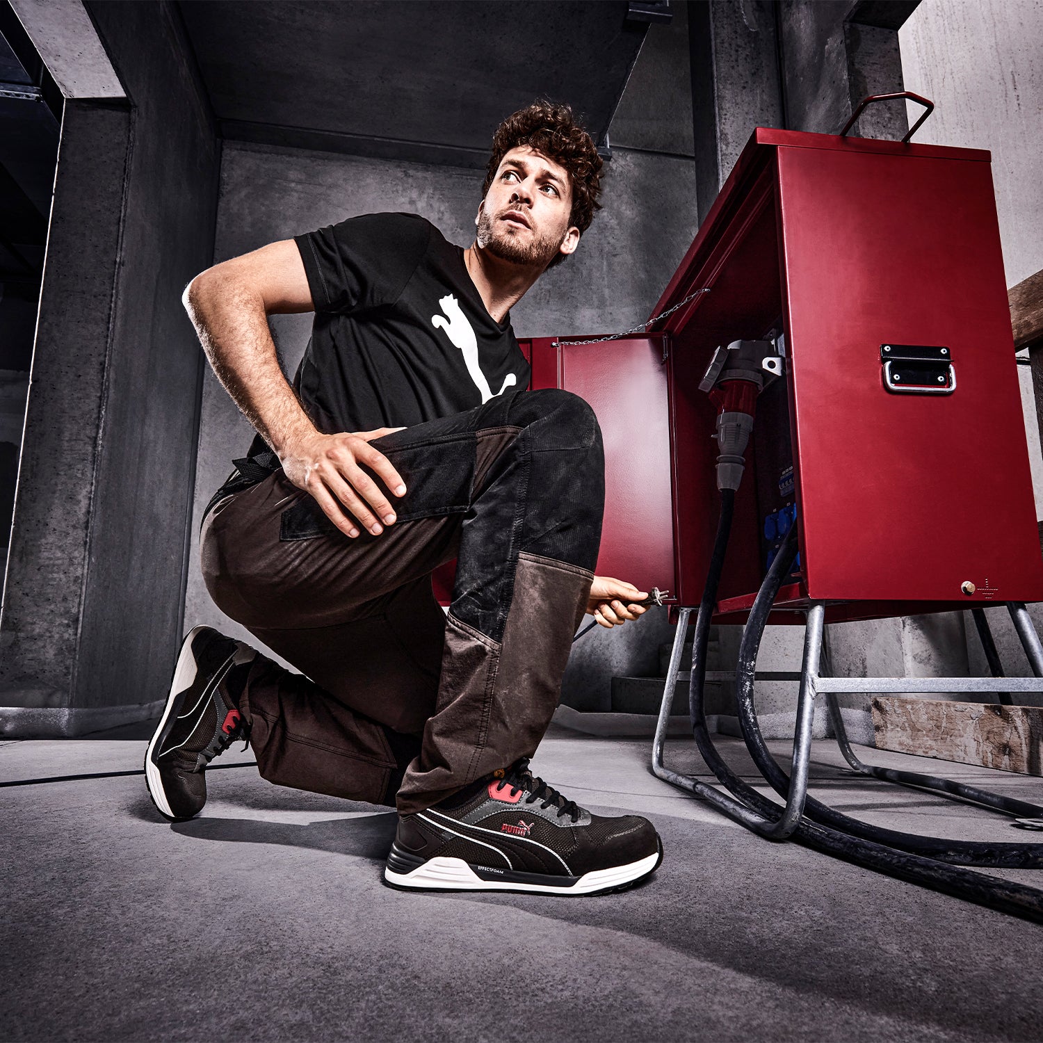 Schwarz-roter PUMA SAFETY FRONTSIDE LOW Sicherheitsschuh S1P ESD mit weißer Sohle und Schnürung, am Fuß getragen von einem Mann in kniender Position vor rotem Schaltschrank.