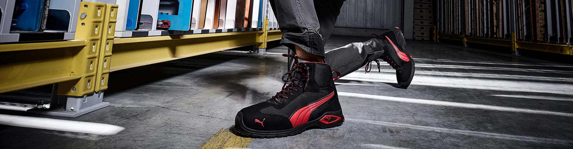Schwarzer Sicherheitsschuh MADRID MID S1PL FO SR von Puma mit roten Akzenten, getragen in einer Lagerhalle auf grauem Industrieboden.