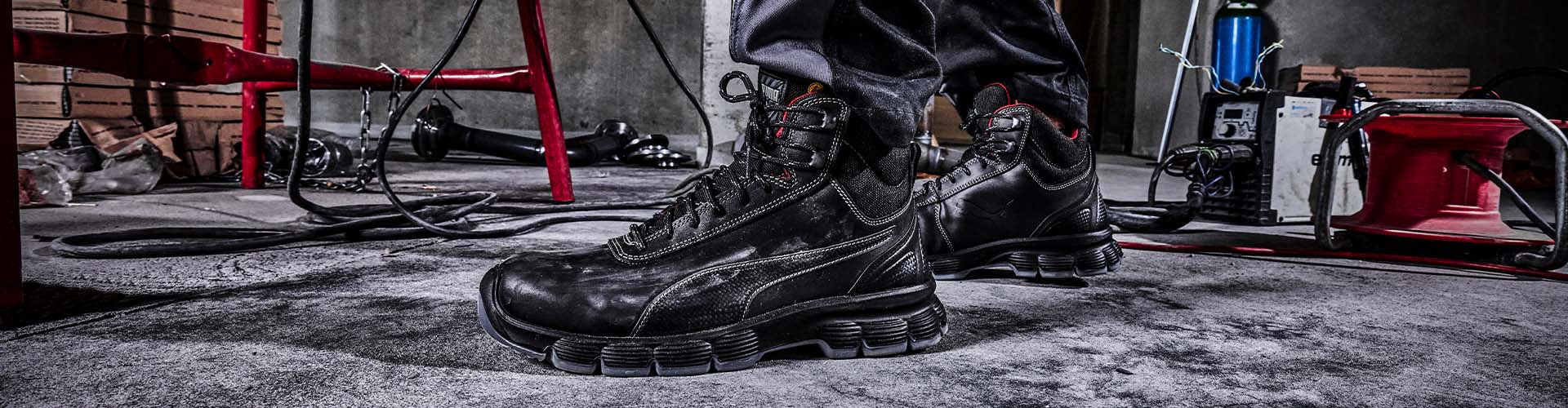 Schwarzer Sicherheitsschuh CONDOR BLACK MID S3L ESD FO SR von Puma, getragen in einer Werkstattumgebung mit Werkzeugen und Kabeln auf Betonboden.