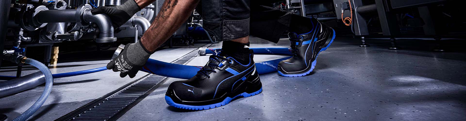 Schwarzer Sicherheitsschuh KRYPTON BLUE LOW S3S ESD FO SR mit blauen Details im Arbeitseinsatz auf Industrieboden, getragen bei der Montage von Rohrleitungen.