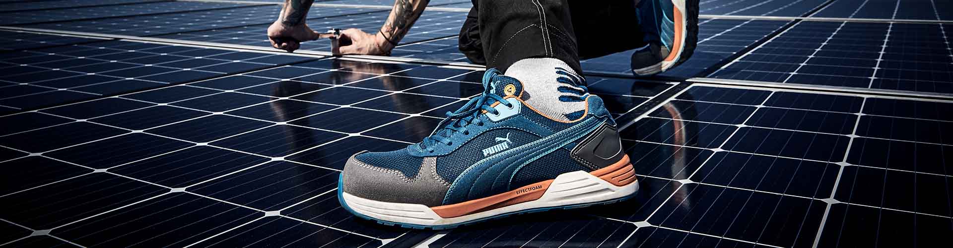 Blau-grauer PUMA SAFETY FRONTSIDE LOW Sicherheitsschuh S1P ESD mit Schnürung und weißer Sohle, am Fuß getragen auf Solarpanelen.