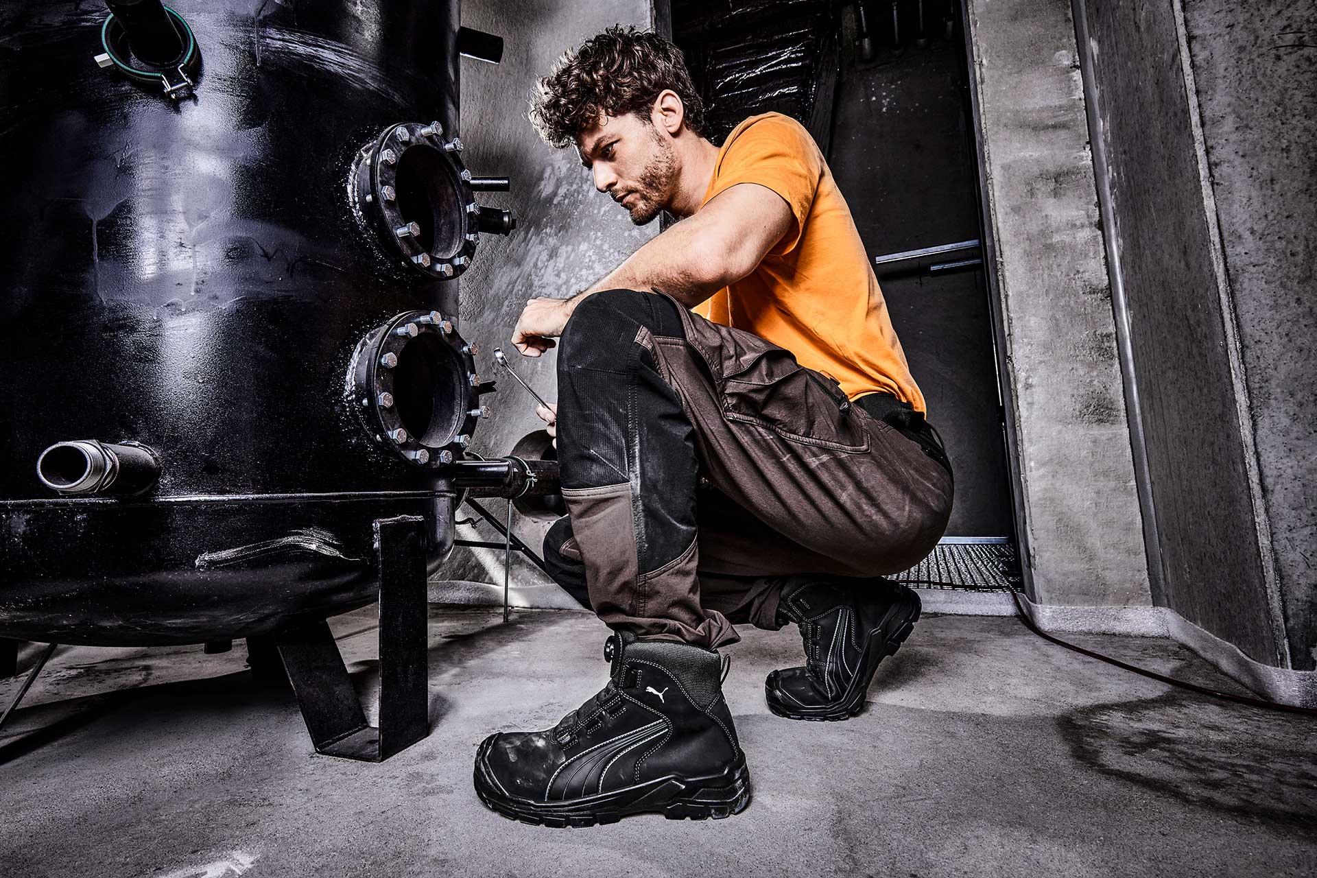 Schwarze PUMA SAFETY CASCADES DISC MID Sicherheitsschuhe S3 mit blauen Akzenten, getragen im Outdoor-Einsatz auf einer schlammigen Baustelle zwischen Steinen