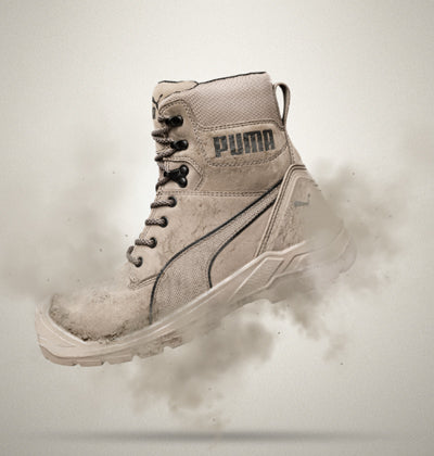 Beiger PUMA SAFETY CONQUEST HIGH Sicherheitsschuh S3 mit hoher Schnürung und robuster Sohle, seitlich freigestellt vor hellem Hintergrund mit aufwirbelndem Staub.
