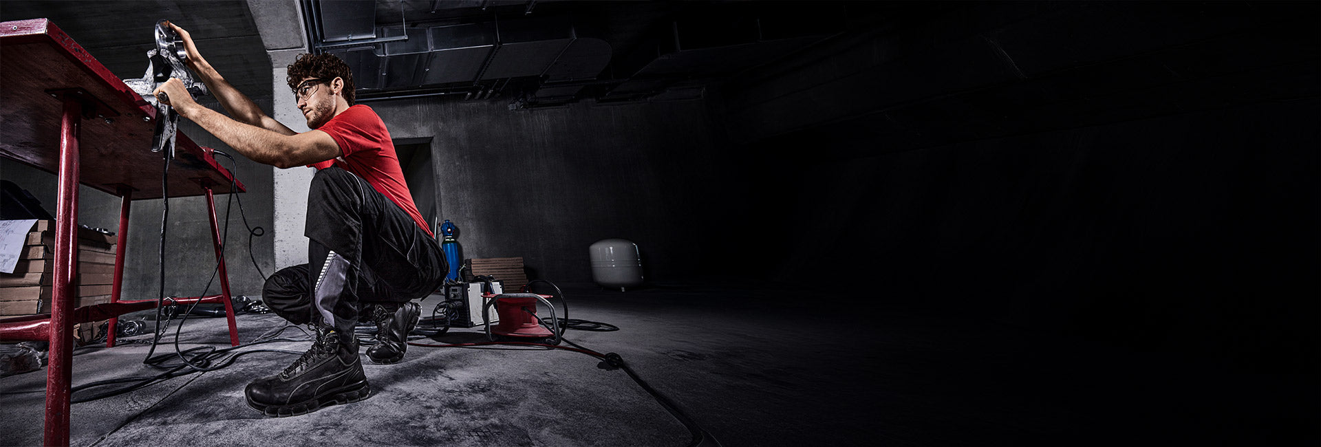 Person wearing black PUMA SAFETY CONDOR BLACK MID safety shoes crouching in an industrial workspace while operating a power tool on a red workbench, surrounded by cables and construction equipment on a concrete floor.