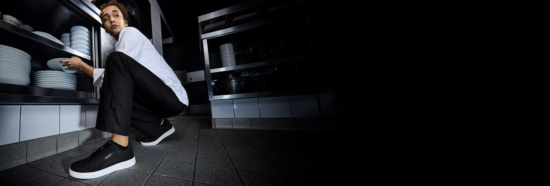 Person in a commercial kitchen crouching to place white plates on a lower stainless-steel shelf, wearing black PUMA SAFETY STELLAR SOFT TOE LOW work shoes with white slip-resistant soles on a tiled floor.