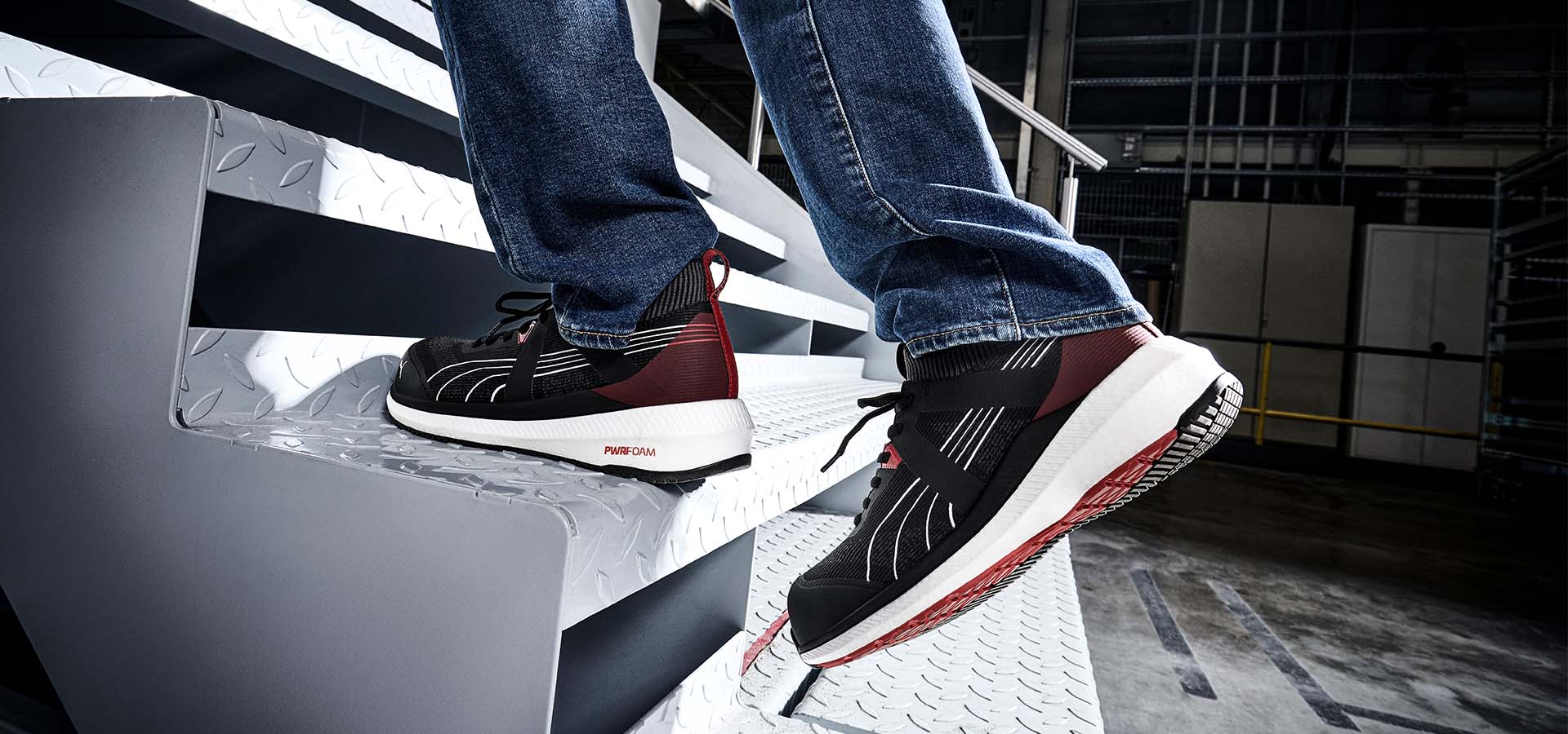 Schwarz-rote PUMA SAFETY TURBO BLK/RED LOW S1PS ESD Sicherheitsschuhe an den Füßen einer Person, die eine Metalltreppe hinaufsteigt. Die Schuhe zeigen ein sportliches Mesh-Design mit weißer Zwischensohle und PWR.FOAM-Dämpfung in einer industriellen Umgebung.