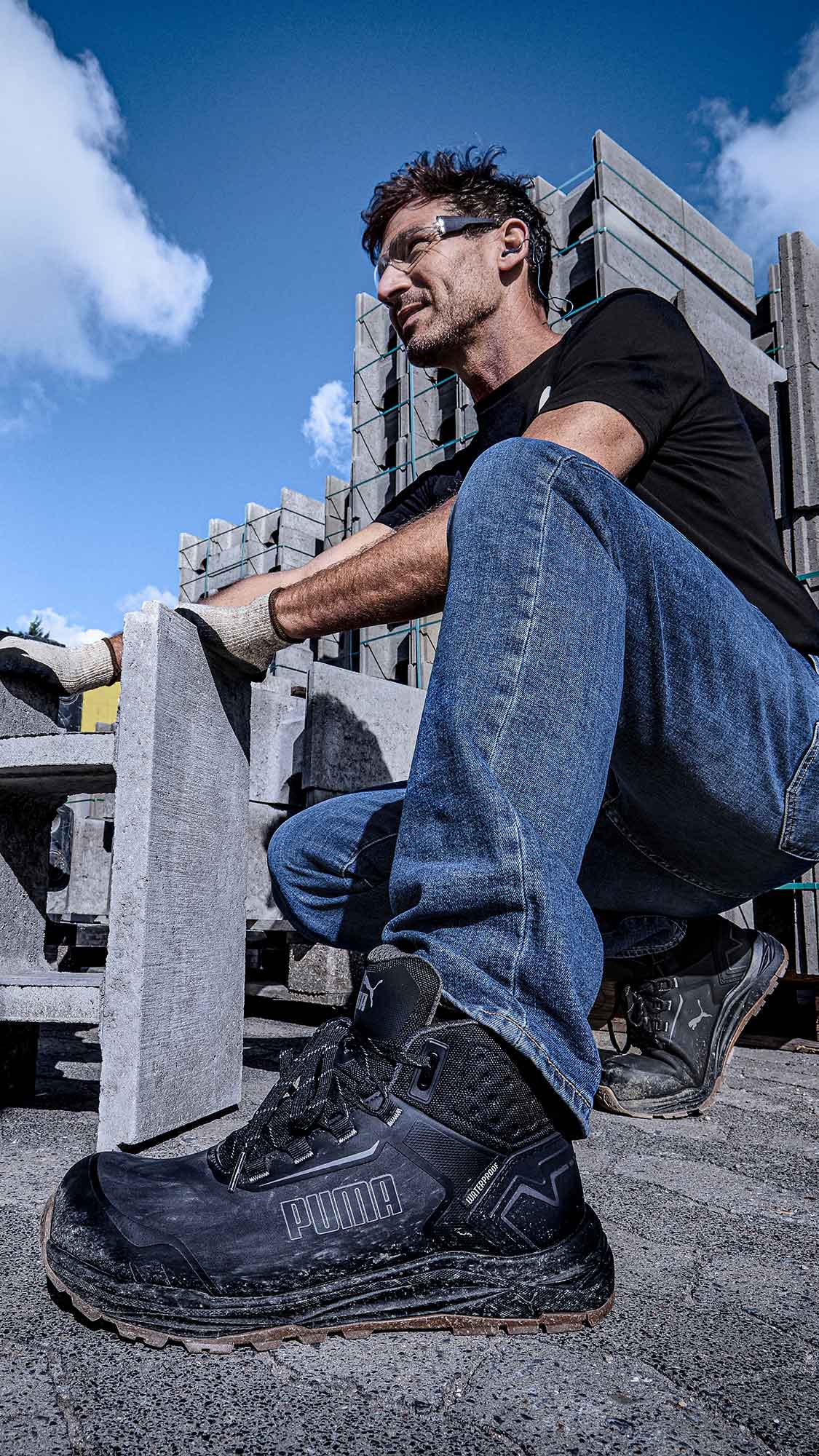 Mobile view: PUMA SAFETY FUSION CTX MID Safety Shoe in Anthracite, worn by a person kneeling on a rough surface next to stacked concrete blocks and a vehicle wheel in an industrial setting
