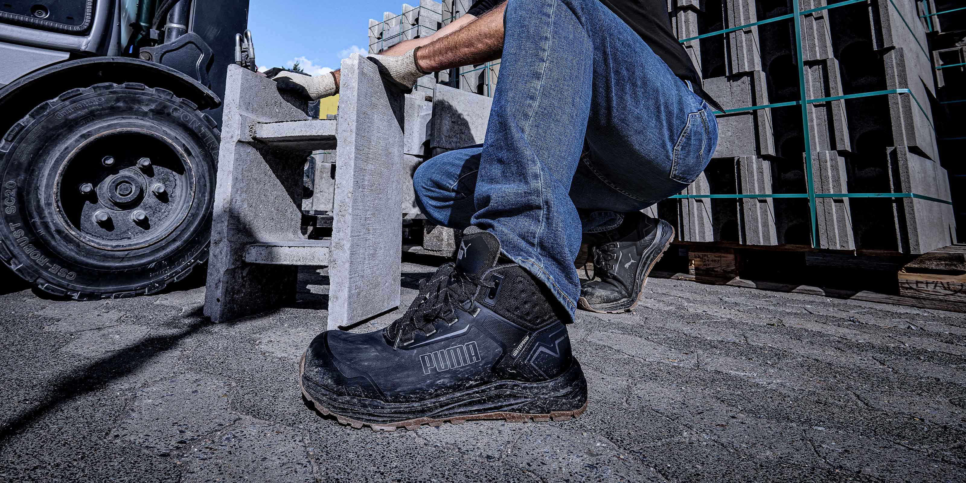 PUMA SAFETY FUSION CTX MID Safety Shoe in Anthracite, worn by a person kneeling on a rough surface next to stacked concrete blocks and a vehicle wheel in an industrial setting