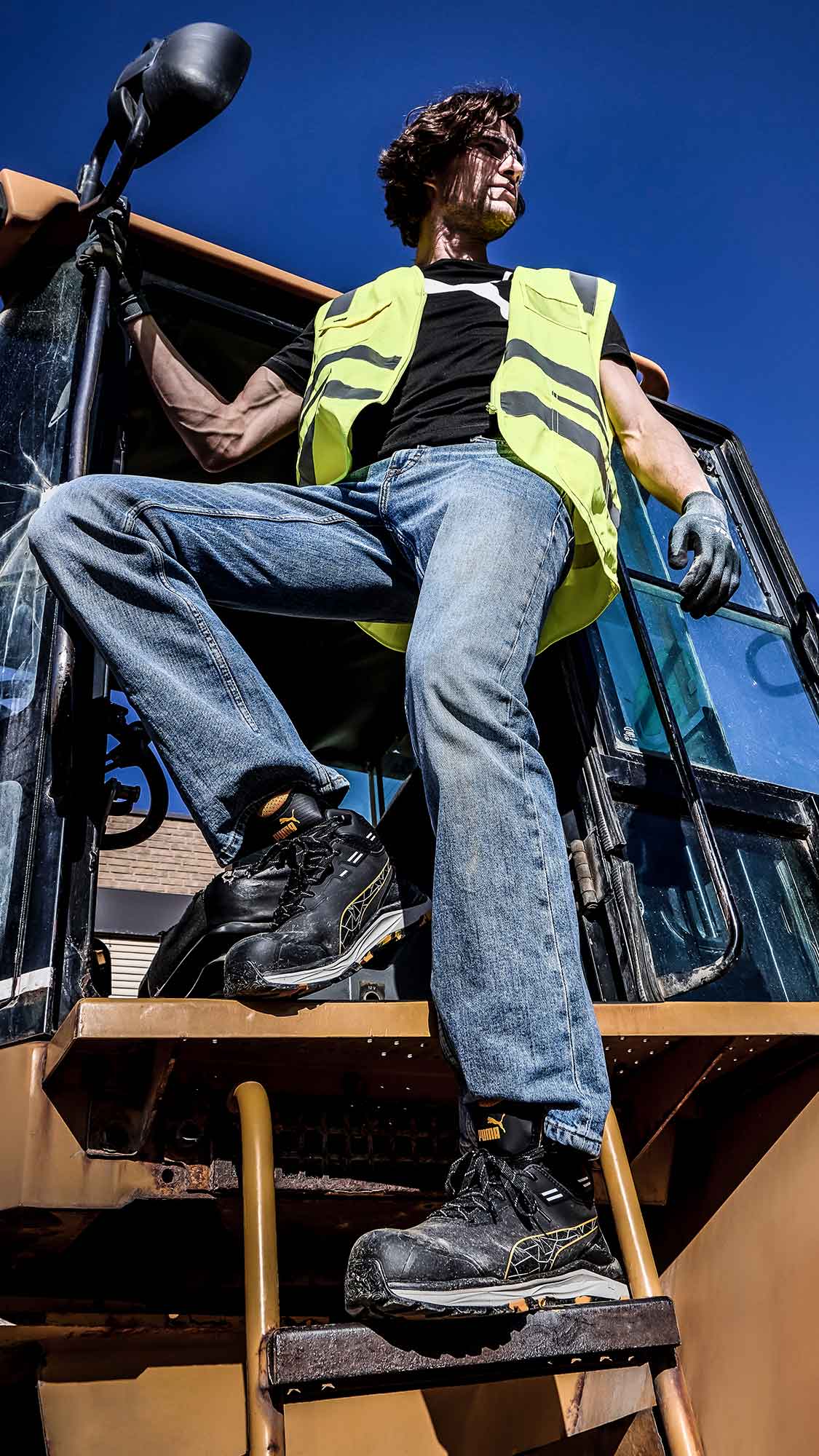 PUMA SAFETY XPLORE MID Sicherheitsschuh S3S ESD in Anthrazit mit gelben Akzenten, getragen von einer Person beim Stehen auf einer Metallleiter an einer Baumaschine im Außenbereich mit blauem Himmel