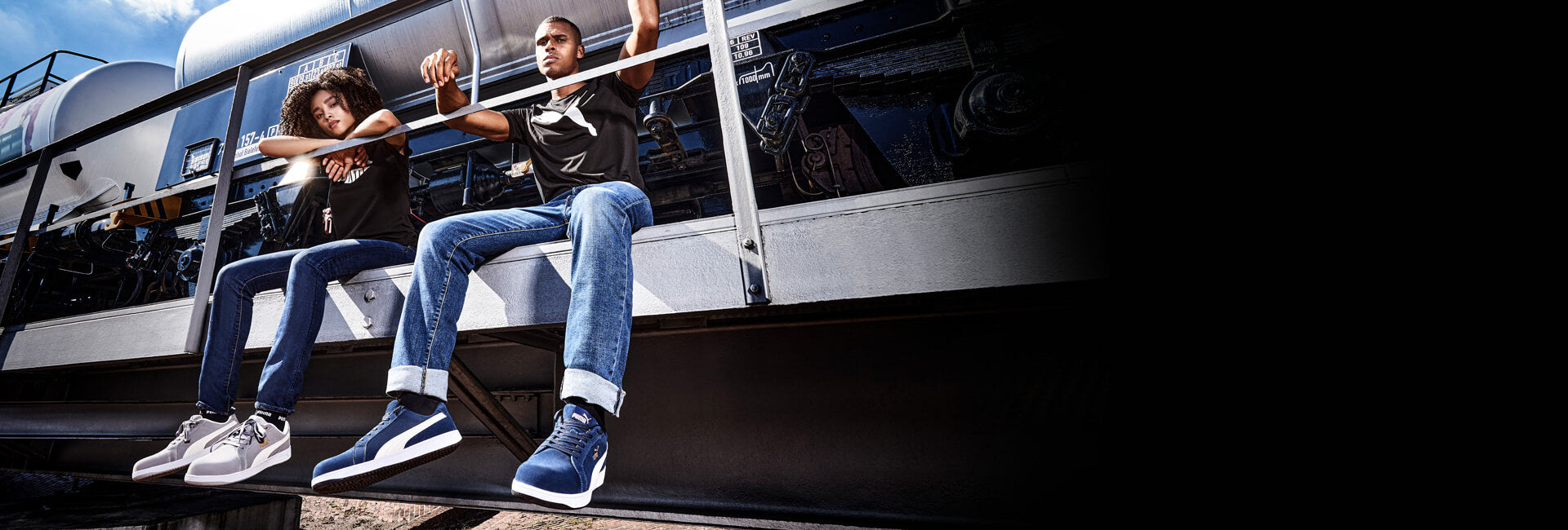 Zwei Personen sitzen auf einer Metallplattform. Sie tragen Jeans, schwarze T-Shirts und Puma Sicherheitsschuhe – links den ICONIC GREY LOW S1PL ESD FO HRO SR in Grau-Weiß, rechts den ICONIC NAVY LOW S1PL ESD FO HRO SR in Blau-Weiß.