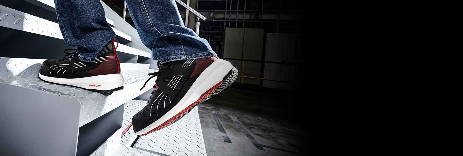 PUMA SAFETY TURBO LOW safety shoe in black with red and white accents, worn by a person climbing a metal staircase in an industrial setting