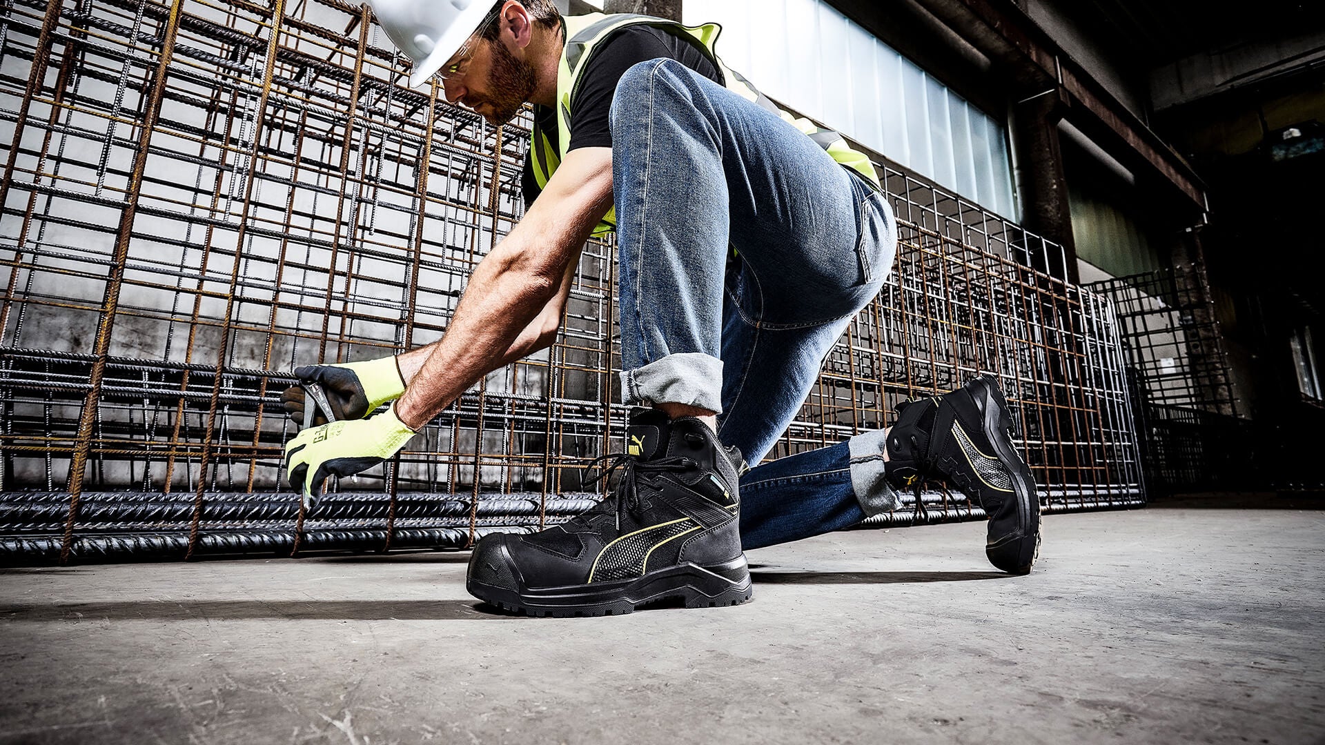 Schwarzer PUMA SAFETY ROCK HD CTX MID Sicherheitsschuh S7S mit gelben Details und robuster Profilsohle, am Fuß getragen von einem Bauarbeiter mit Schutzhelm und Handschuhen beim Arbeiten an Stahlgittern.